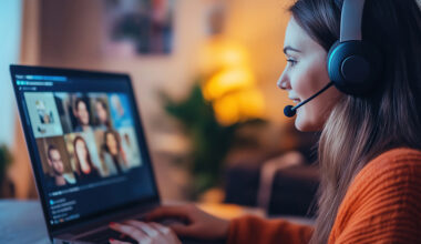 Photo of young caucasian woman working at her desk at home wearing a headsset and engaged in a videoteleconferencing call on her laptop computer.