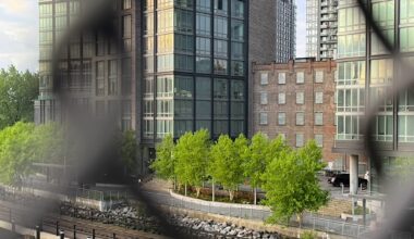 View of modern buildings alongside a waterfront park, framed by a fence, with greenery in the foreground.