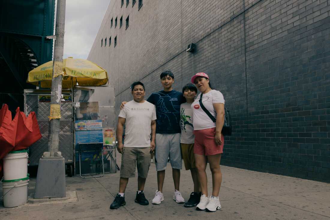 Juan David Iza, center-right, and his mother Lida Iza, right, pose for a portrait with their family along Roosevelt Avenue and 99 Street, as residents navigate the bustling avenue in Queens, New York, U.S., on Sunday, June 7, 2026. CREDIT: José A. Alvarado Jr. for NPR, @josealvarado