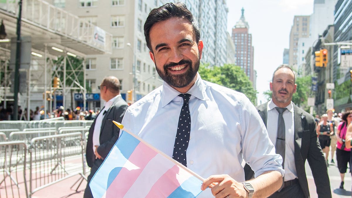 Zohran Mamdani smiles while holding a transgender pride flag in 2025