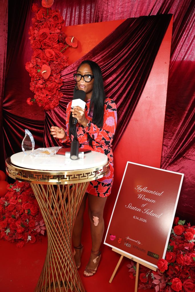 A woman in a red floral dress stands at a podium, speaking into a microphone. Behind her is a red curtain and a sign that reads "Influential Women Staten Island 6.4.2026".