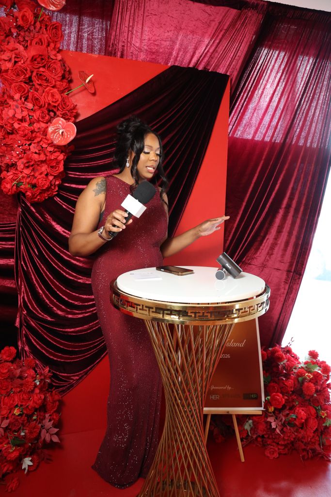 A woman in a red sequined dress standing in front of a red and black backdrop, holding a microphone and gesturing with her hand. The backdrop features red curtains and a large drum.