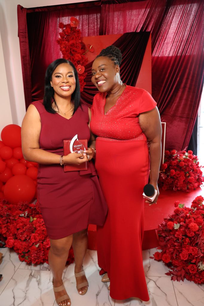 Two women in red dresses standing in front of a red floral backdrop, one holding an award.