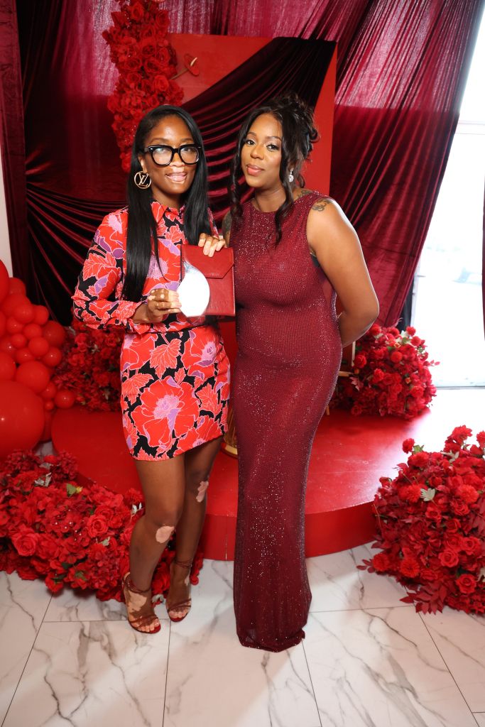 Two women in formal dresses posing in front of a red floral backdrop, one wearing a red floral dress and the other a red sequined gown.