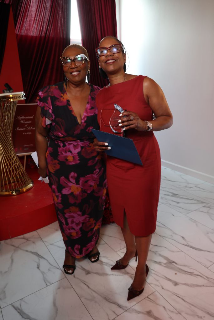 Two women in formal attire, one in a red dress and the other in a floral print dress, holding an award and smiling.