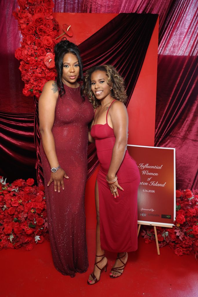 Two women in red dresses posing in front of a red and black backdrop with floral arrangements and a sign that says "Influential Women of Staten Island 6.14.2026".