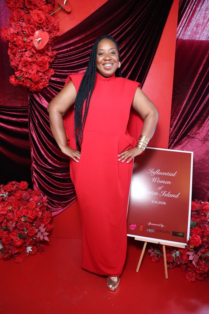 A smiling woman wearing a red dress stands in front of a backdrop of red roses and velvet curtains. A sign behind her reads "Influential Women of Staten Island 6.14.2026".