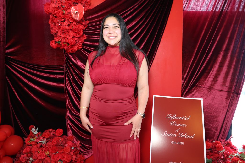 A smiling woman in a red dress stands in front of a red velvet backdrop, holding a sign that reads "Influential Women of Staten Island 6.14.2026".