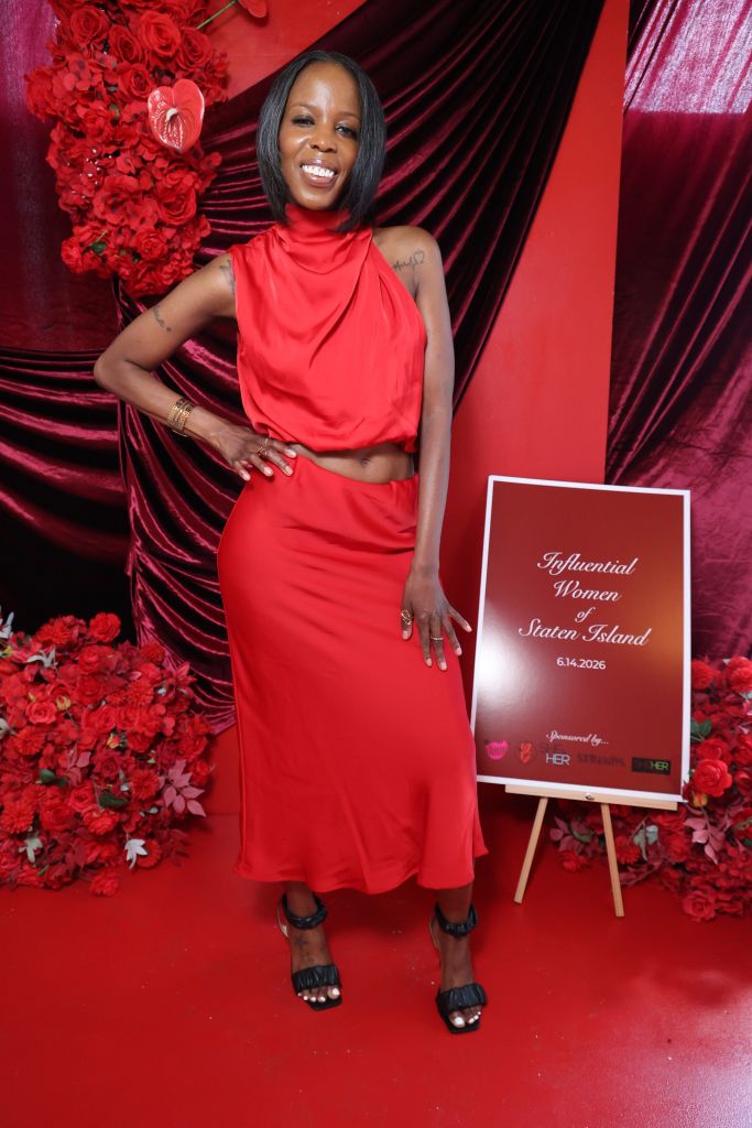 A smiling woman in a red dress stands in front of a red floral backdrop, holding a sign that reads "Influential Women of Staten Island 6.14.2026".