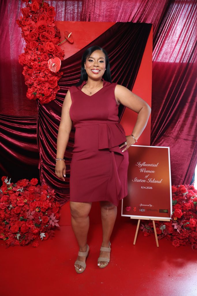 A woman in a red dress stands in front of a red and black backdrop holding a sign that says "Influential Women of Staten Island 6.14.2026".