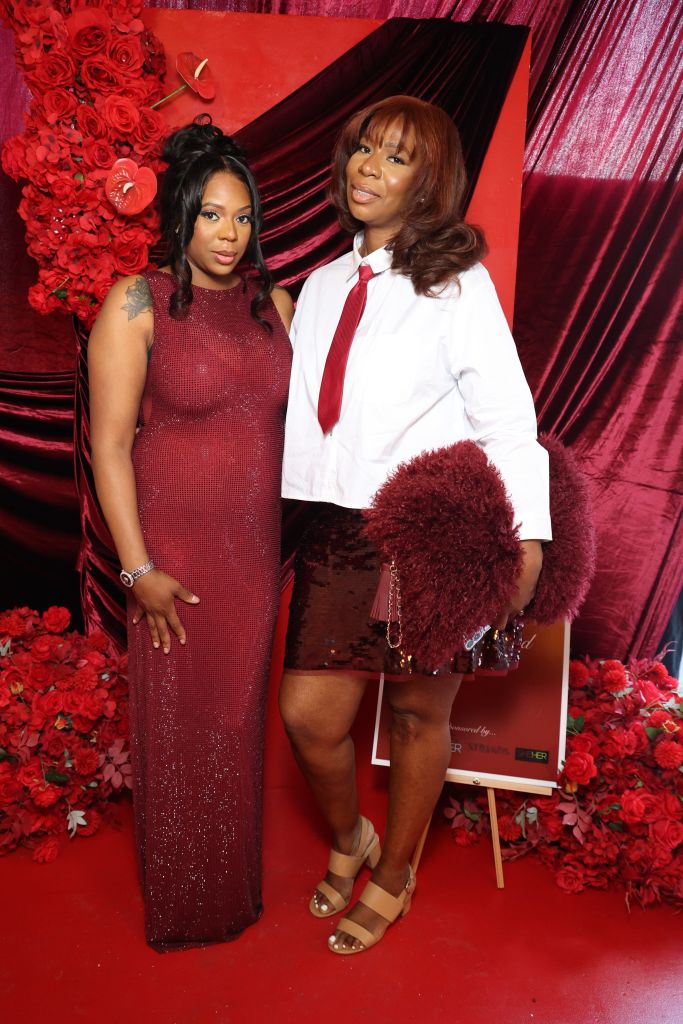 Two women posing in front of a red floral backdrop, one wearing a red sequin dress and the other a white suit with a red tie.