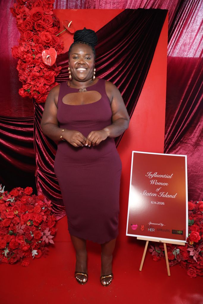 A smiling woman in a burgundy dress stands in front of a red floral backdrop, holding a sign that reads "Influential Women Staten Island 6.14.2026".