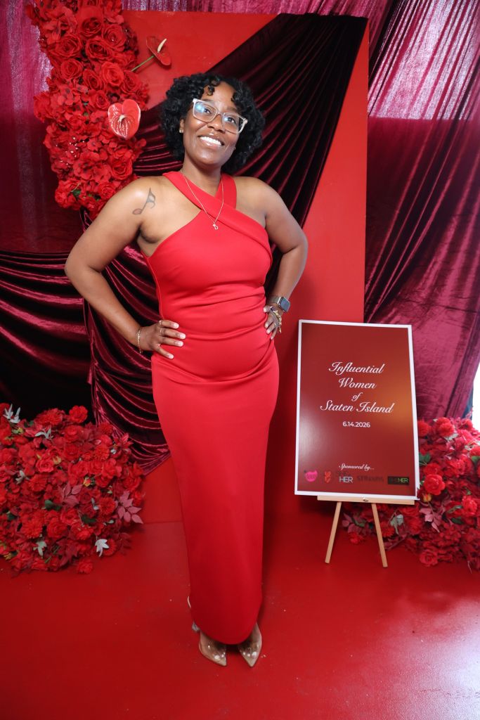 A smiling woman in a red dress stands in front of a red and black backdrop, holding a sign that says "Influential Women of Staten Island 6.14.2026".
