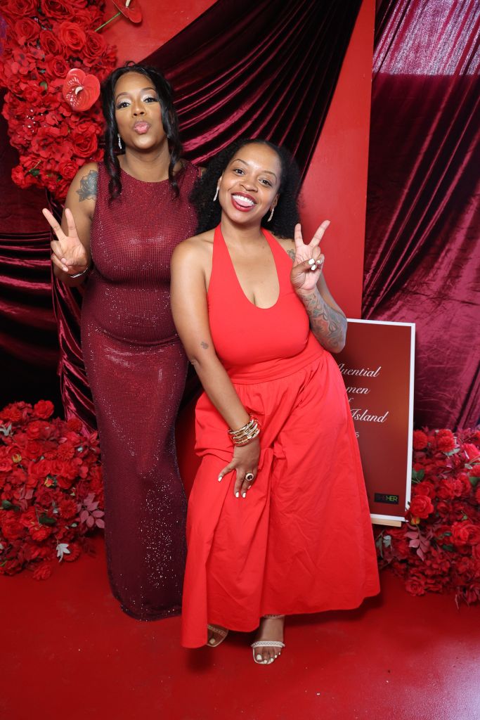 Two women in red dresses posing and making peace signs in front of a red floral backdrop with a sign that says "Influential Women of Long Island".