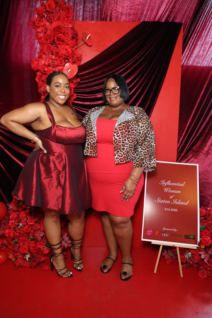 Two women in red dresses posing in front of a red and black backdrop, holding a sign that says "Influential Women of Staten Island 6.14.2026".