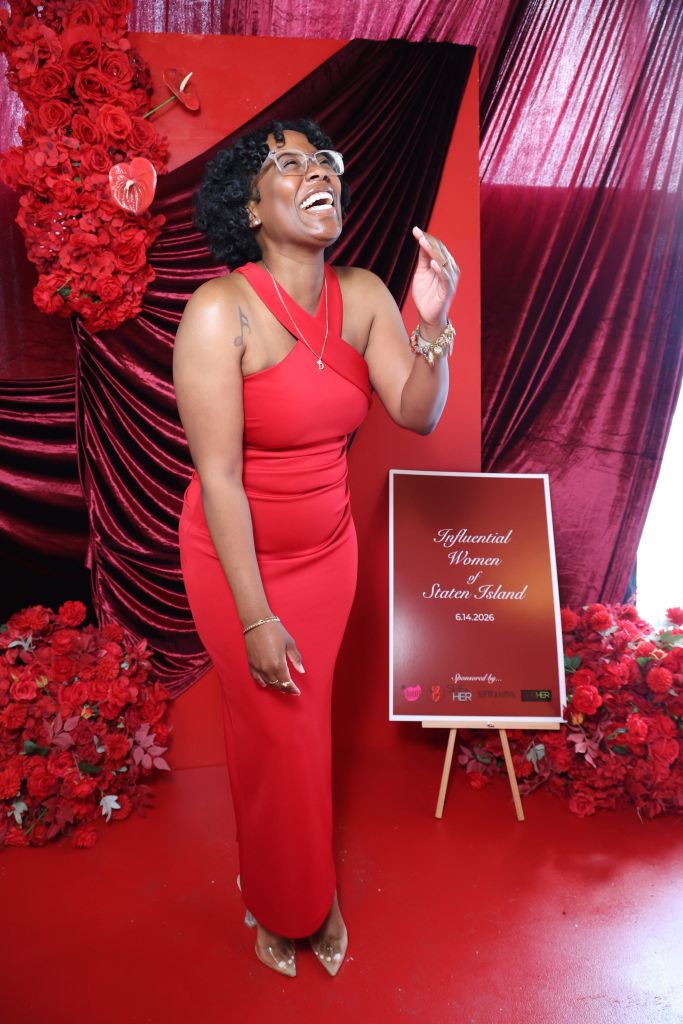 A smiling woman in a red dress stands in front of a red and black backdrop, holding a sign that reads "Influential Women of Staten Island 6.14.2026".