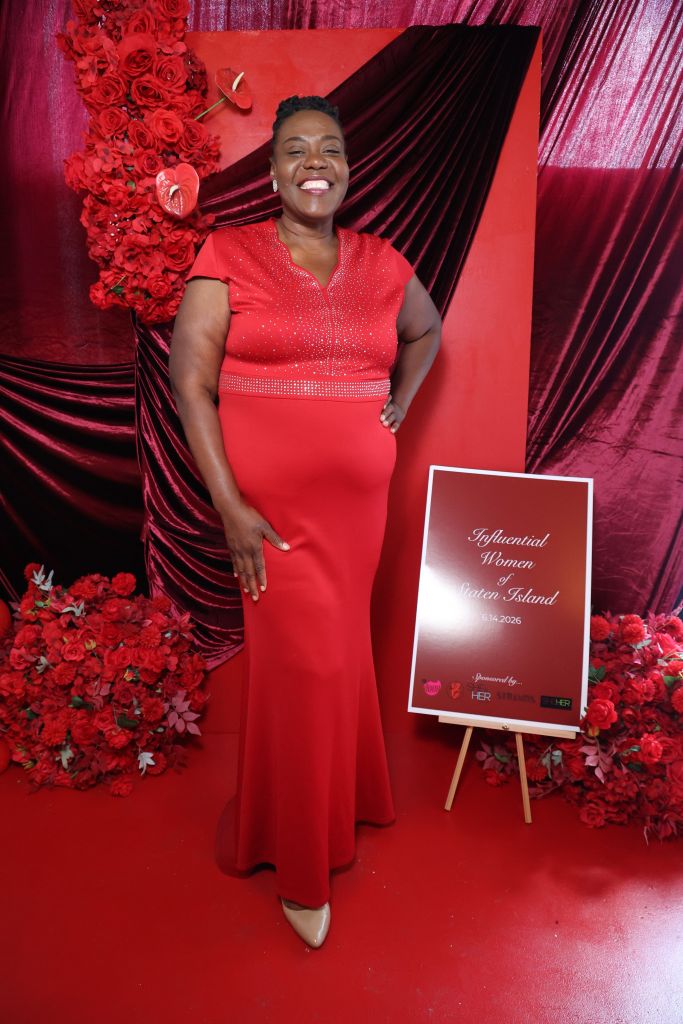A smiling woman in a red sequined dress stands in front of a red and black backdrop, holding a sign that reads "Influential Women of Staten Island".