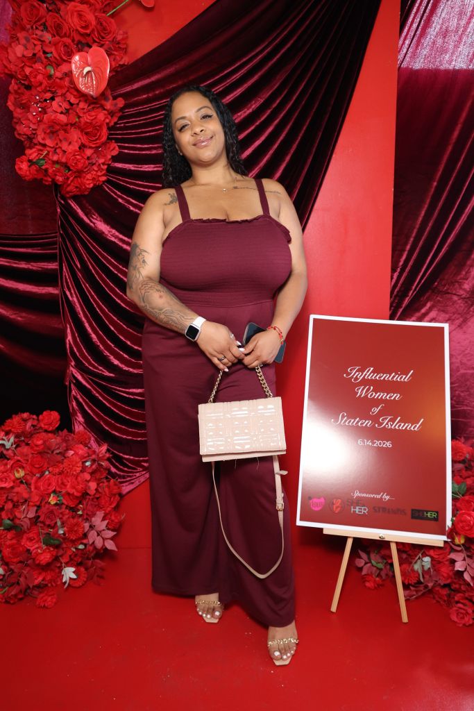 A woman in a burgundy dress stands in front of a red velvet backdrop, holding a pink clutch and a sign that says "Influential Women of Staten Island 6.14.2026".