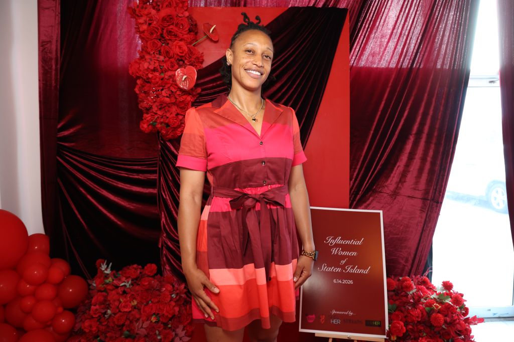A smiling woman wearing a red and pink dress stands in front of a red and black backdrop with a sign that says "Influential Women of Staten Island 6.14.2006".