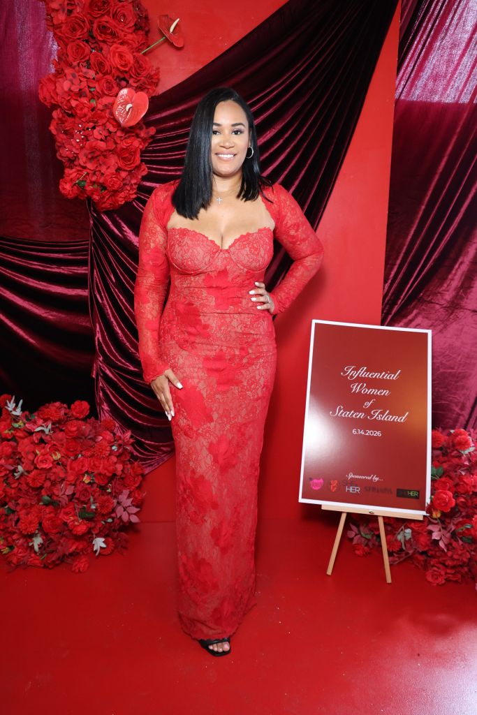 A woman in a red lace dress stands in front of a backdrop of red roses and velvet curtains, holding a sign that reads "Influential Women of Staten Island 6.14.2026".