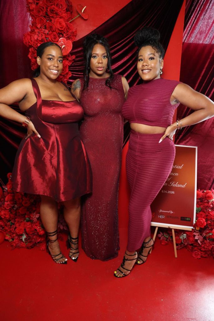 Three women in red dresses posing in front of a red floral backdrop with a sign that says "Virtual Women's Summit on Island 6.14.2026".