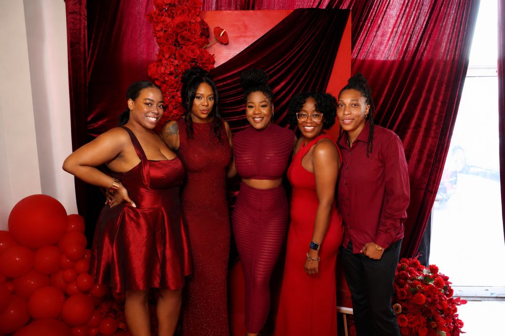 A group of five young women wearing red dresses and tops posing together in front of a red and black backdrop with floral arrangements.
