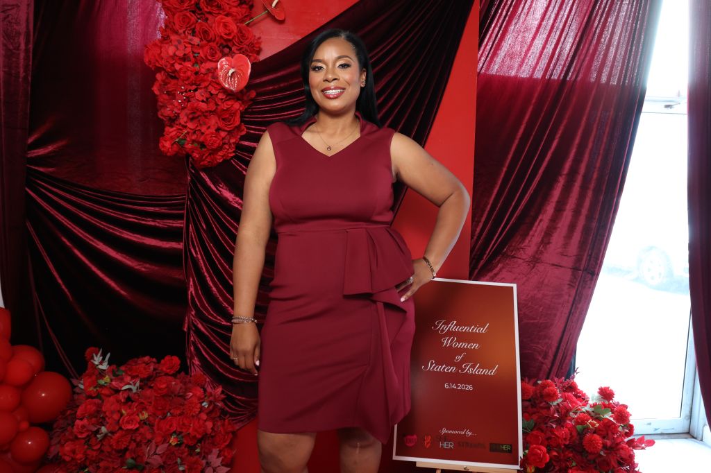 A smiling woman in a red dress stands in front of a red and black backdrop, holding a sign that reads "Influential Women of Staten Island 6.14.2023".