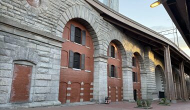Historical brick and stone archway structure with windows, situated alongside a paved walkway.