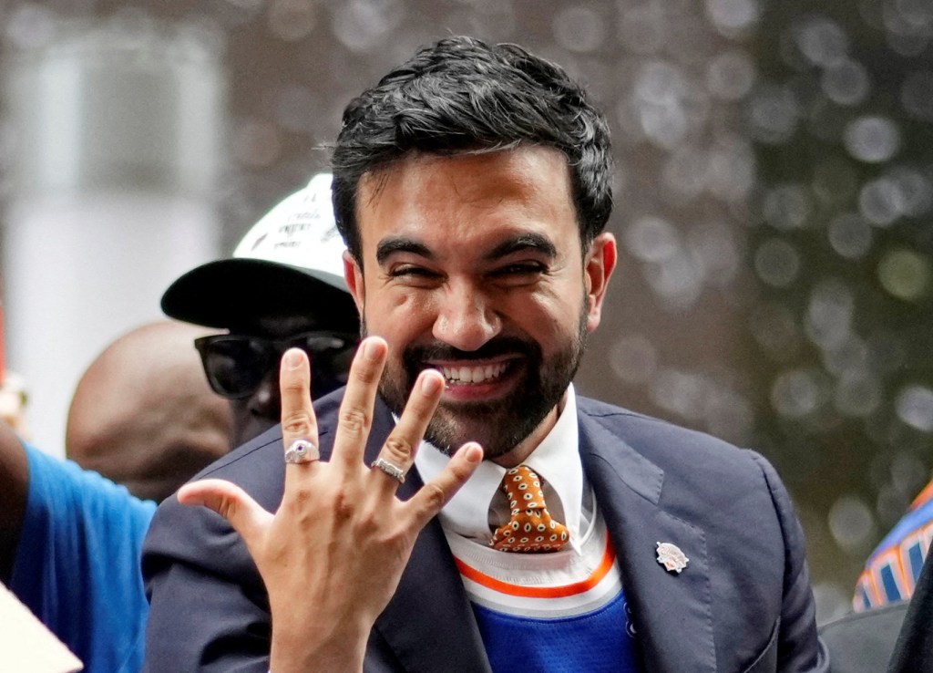 New York City Mayor Zohran Mamdani smiling and holding up four fingers at the NBA New York Knicks parade.