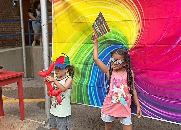 Kids posed in front of the rainbow swirl tapestry, one showing off a new balloon-animal dog, and another holding a mini Juneteenth flag high.