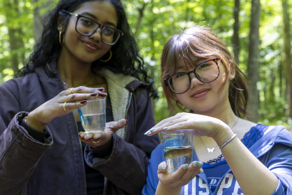 Two students pose together holding trout fingerlings in a cup, poised to be released into a stream