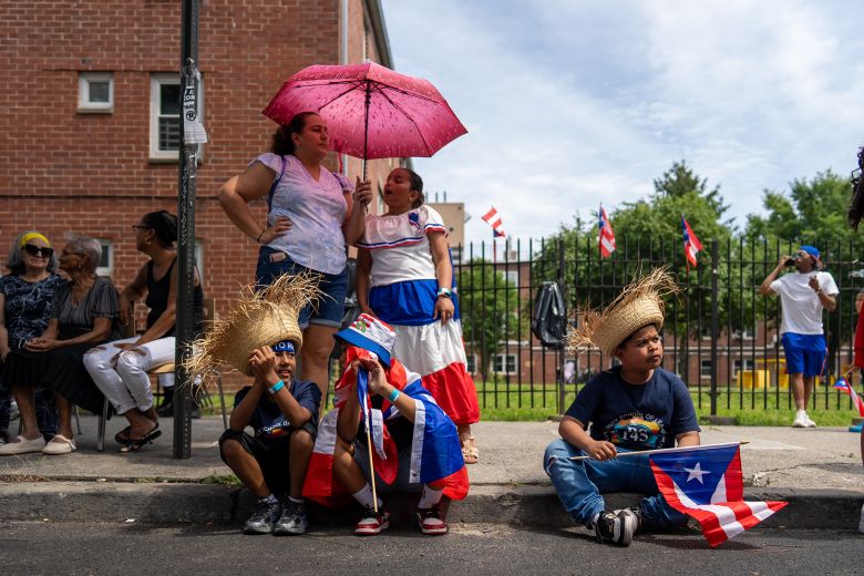 A family shares an umbrella to beat the New York heat while two boys wear traditional pava straw hats.
