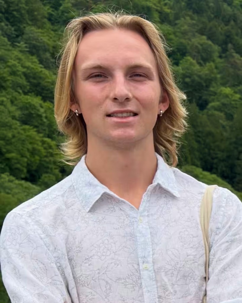 A young man with blonde hair smiles at the camera, wearing a white patterned shirt.