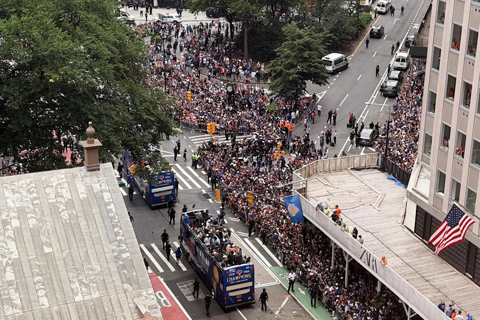 Crowds gather on a city street for a parade, featuring floats with "Champions" banners. Police manage the event, and a U.S. flag is visible