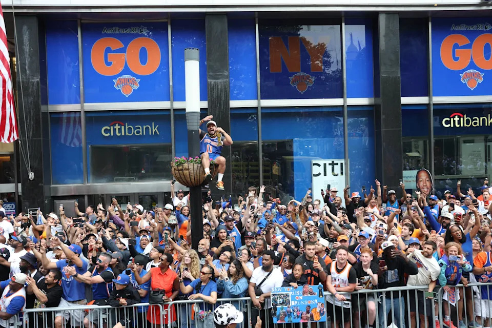 Large crowd celebrates outside Citibank with New York sports team signs, including a person elevated in the center