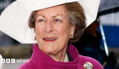 Lady Pamela Hicks in a pink coat with a gold brooch on the shoulder, and a large white hat.