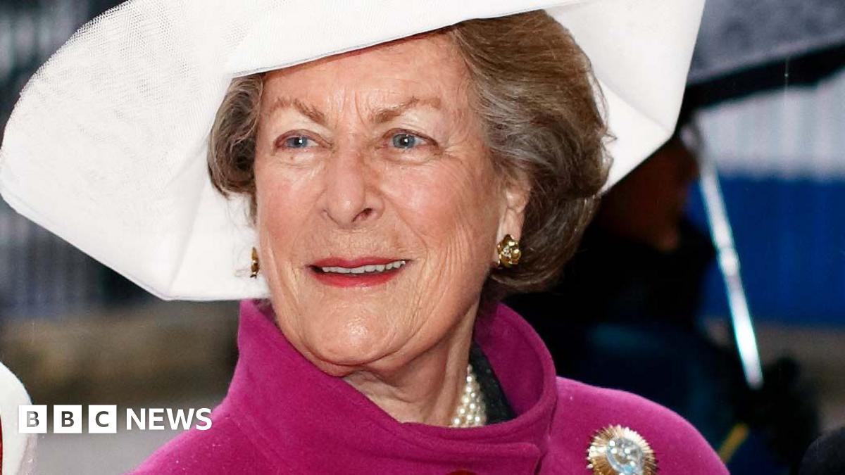 Lady Pamela Hicks in a pink coat with a gold brooch on the shoulder, and a large white hat.