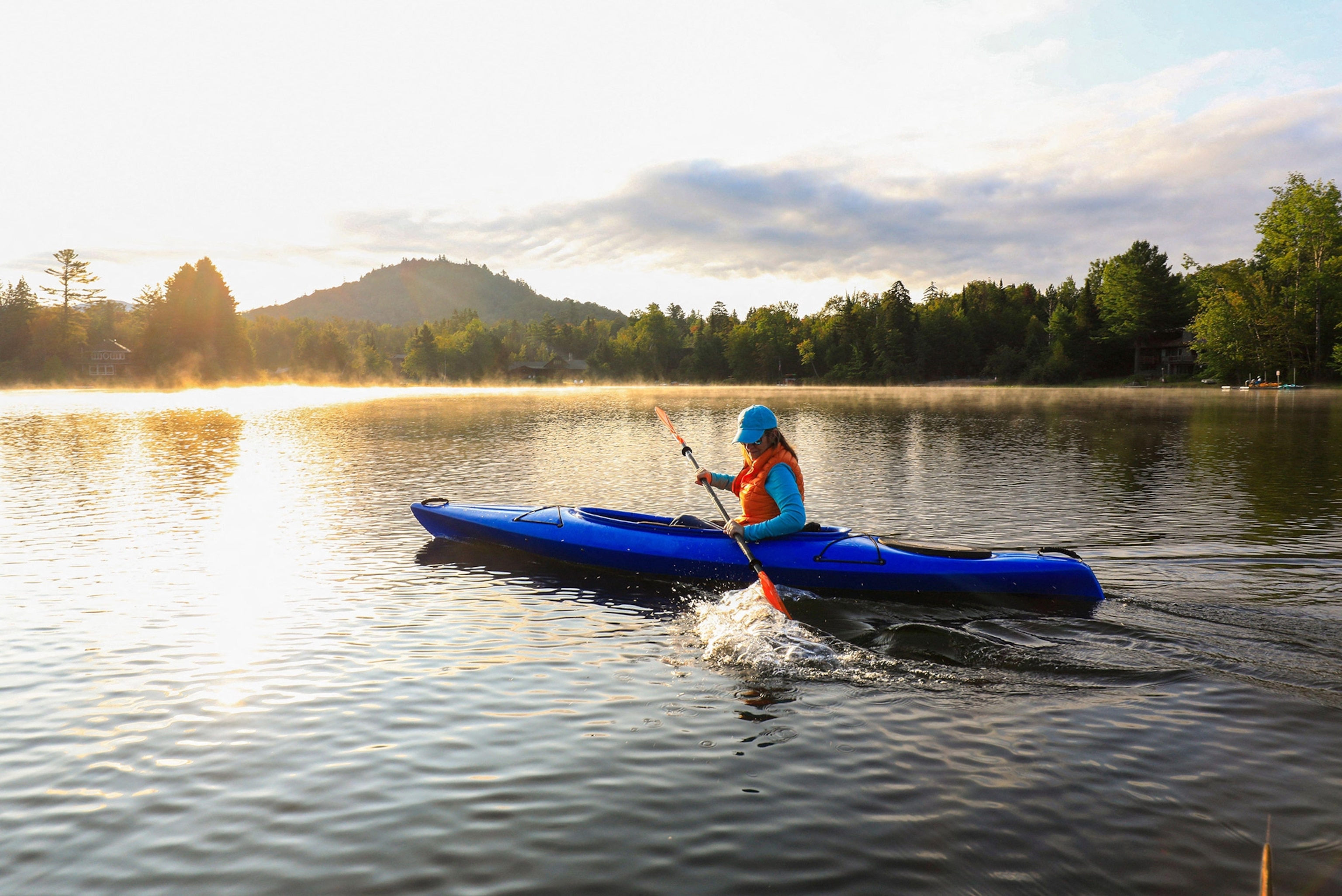 A woman in a blue kayak rows across a glassy lake with mountains and woods behind her and a sunburst overhead