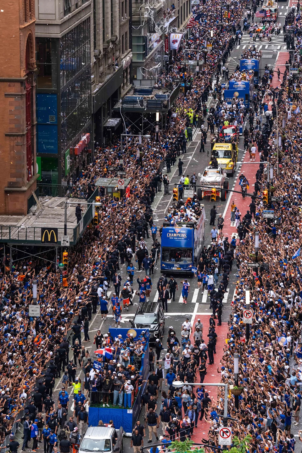 Aerial view of a large city parade with crowds lining the street, police presence, and buses transporting a sports team