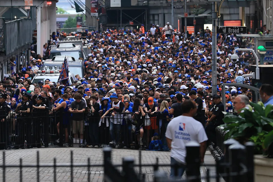 Large crowd gathered on a city street for a public event, with some wearing sports jerseys and caps. Police and event staff are managing the scene