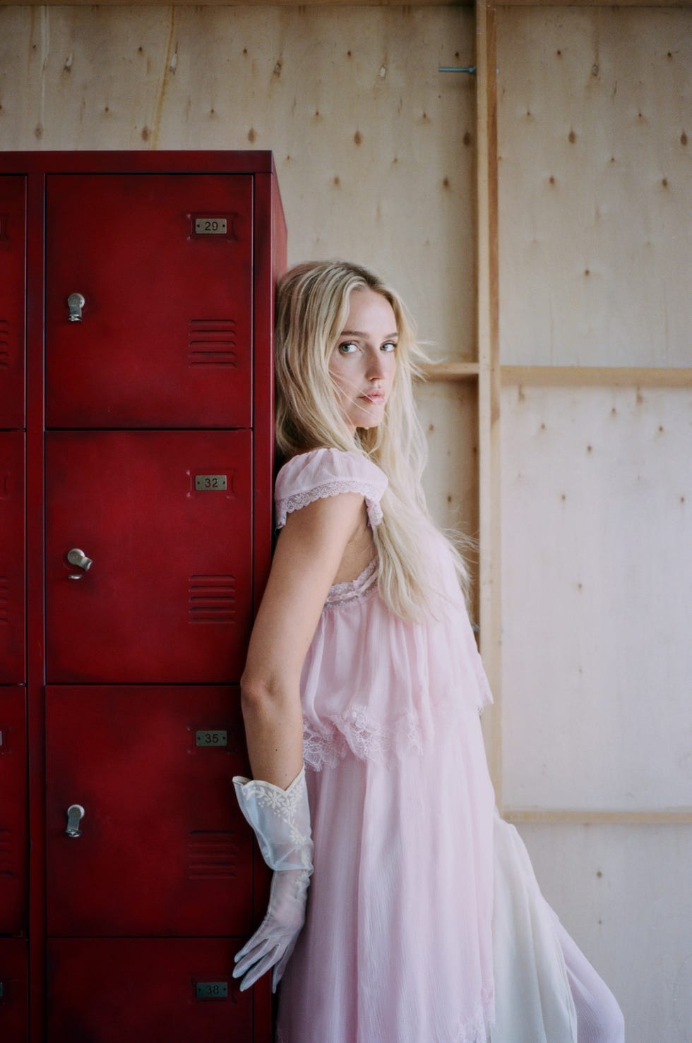 Person in a pink dress and white gloves leaning against red lockers.