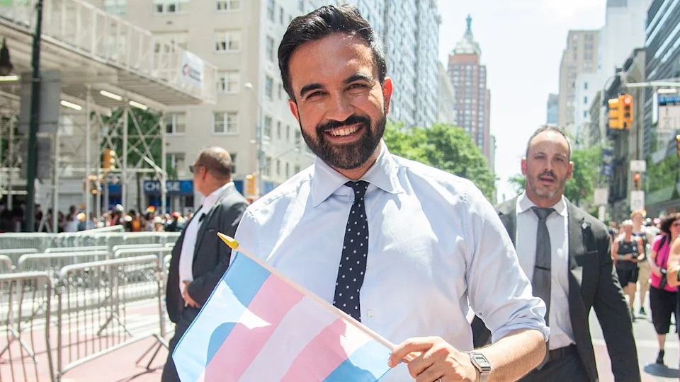 Zohran Mamdani smiles while holding a transgender pride flag in 2025