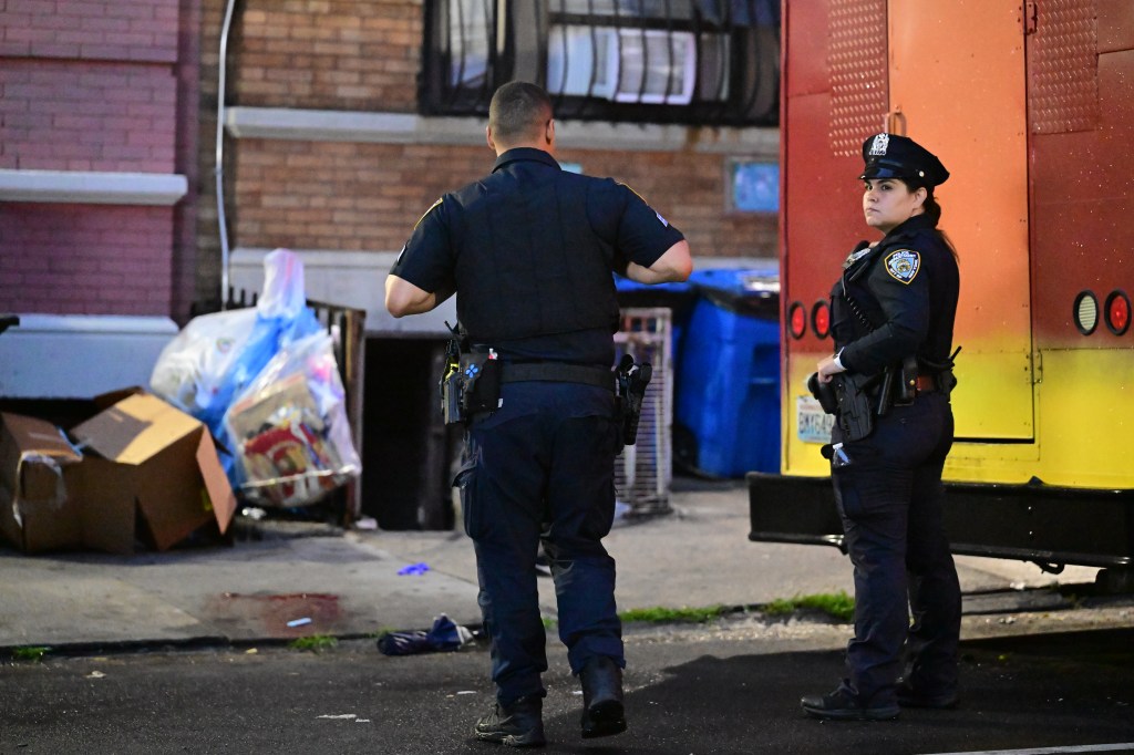 Two police officers stand on a street, with one facing away and the other looking towards the right, near a building and a yellow vehicle.