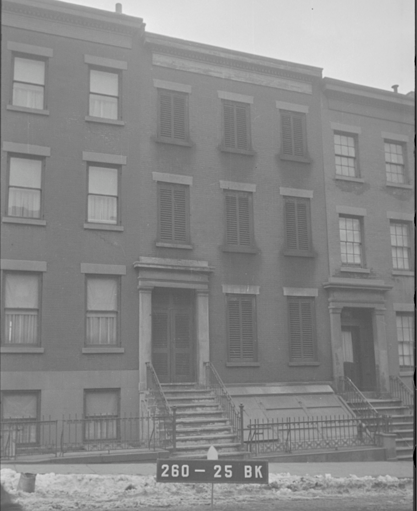 A black and white image of a multi-story brick building with multiple windows and a front entrance featuring stairs. A sign with the numbers '260 - 25 BK' is visible in the foreground, alongside a snow-covered sidewalk.