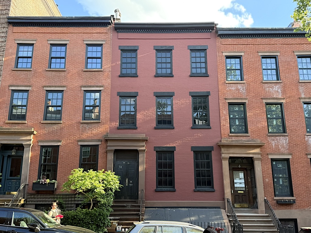 A row of three historical brownstone buildings in a residential area, featuring large black-framed windows and varying wall colors, with one building in a light red hue. Stairs lead up to the entrances, and greenery is present at the base.