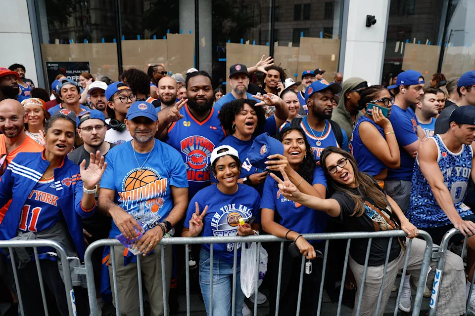 A crowd of diverse, enthusiastic sports fans gathered behind a barrier outside, many wearing team jerseys and hats, posing and smiling for the camera