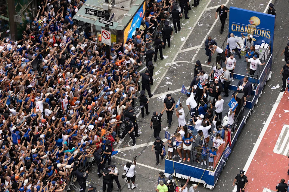 Crowd gathers around a parade float with people celebrating a championship victory, escorted by police on a city street