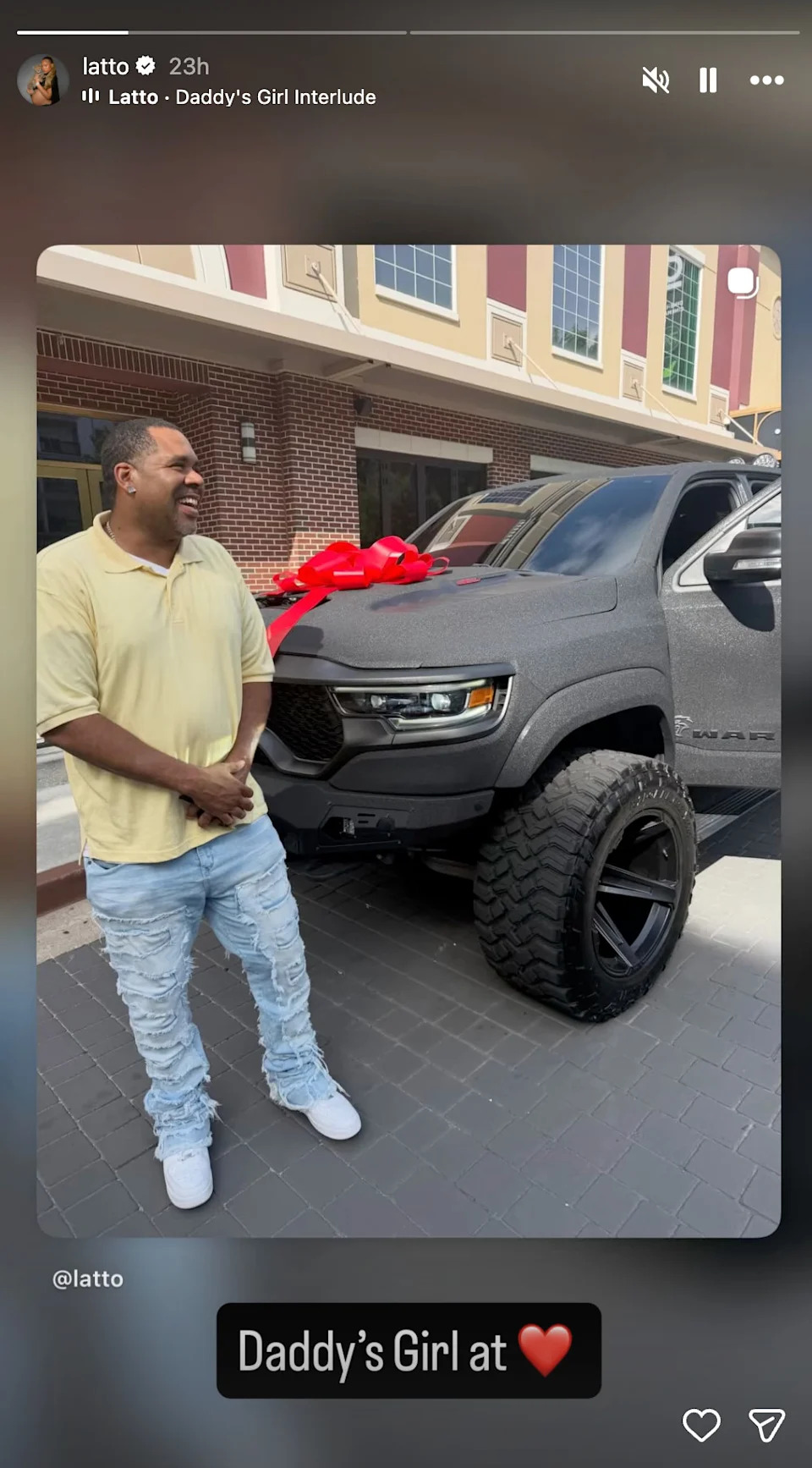 A man smiles next to a large black truck with a red bow on the hood, standing in front of a building.