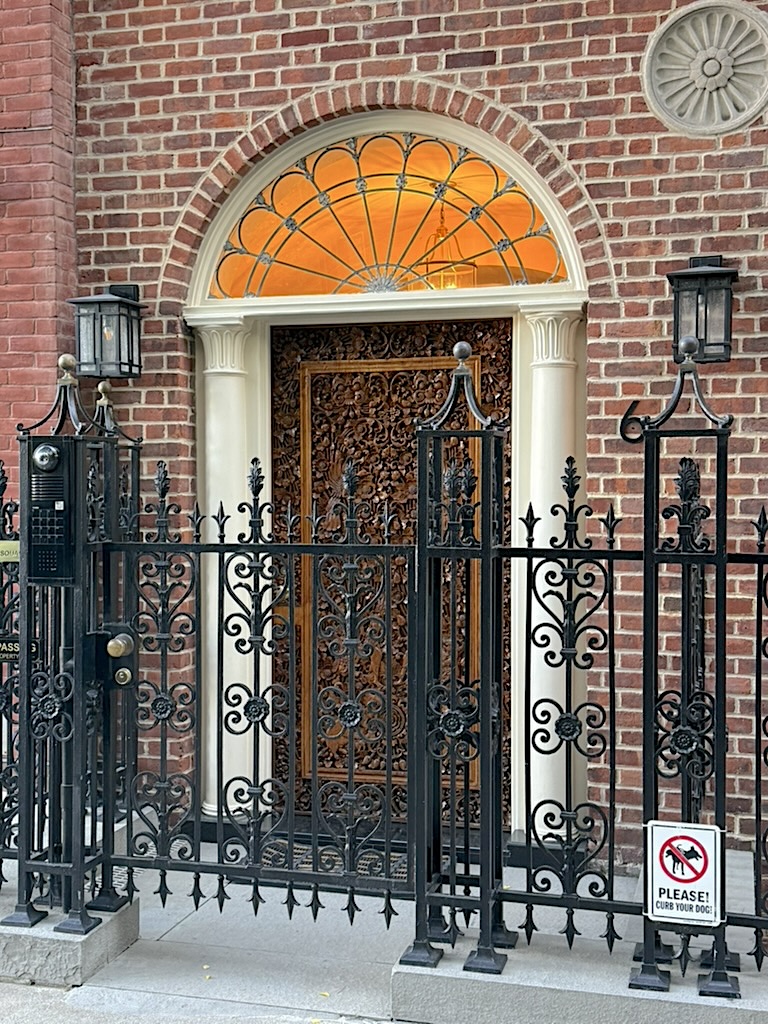 An ornate door with intricate wood detailing, framed by a semi-circular window above, situated behind a black wrought-iron fence in front of a brick wall.