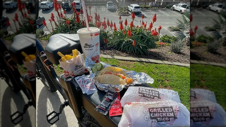 A feast of food. including tacos, crinkle cut fries, hot sauce, and a drink, from Del Taco on a bicycle at the beach.